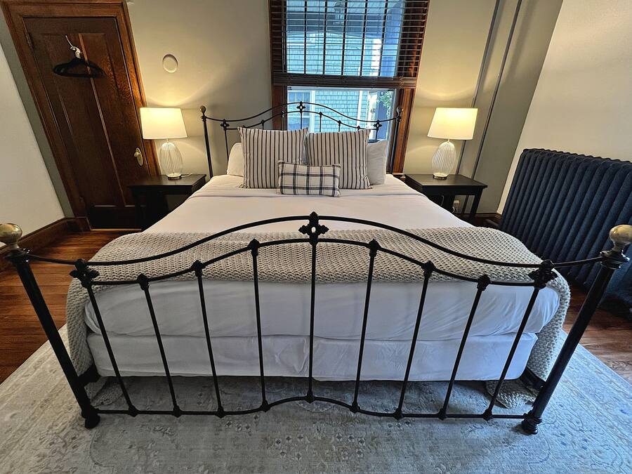 A neatly arranged bedroom with a metal bed frame, a large window with blinds, two bedside tables with lamps, and a radiator.