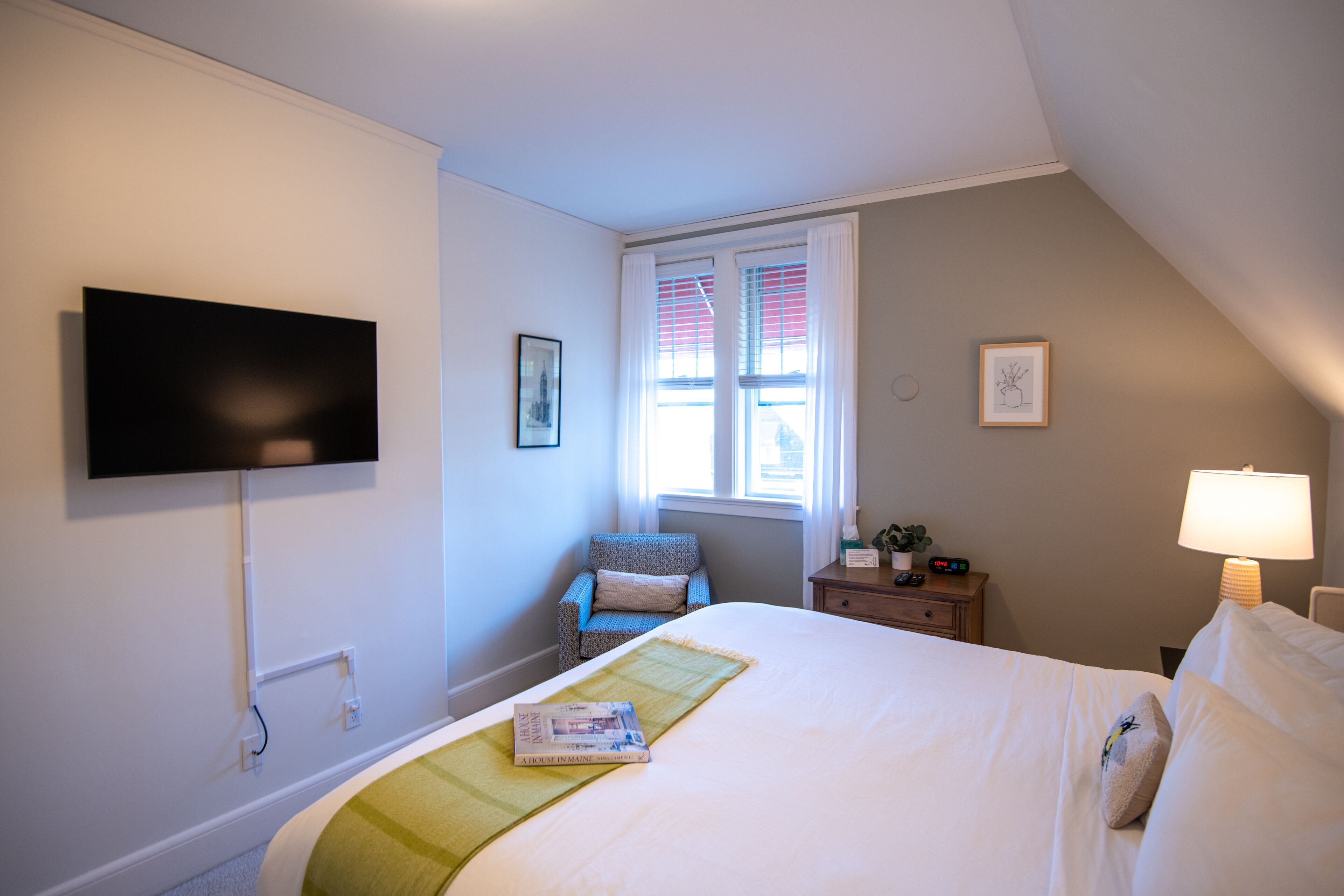 A hotel room with a bed, a TV mounted on the wall, a chair, a nightstand with a lamp, and a window with curtains.