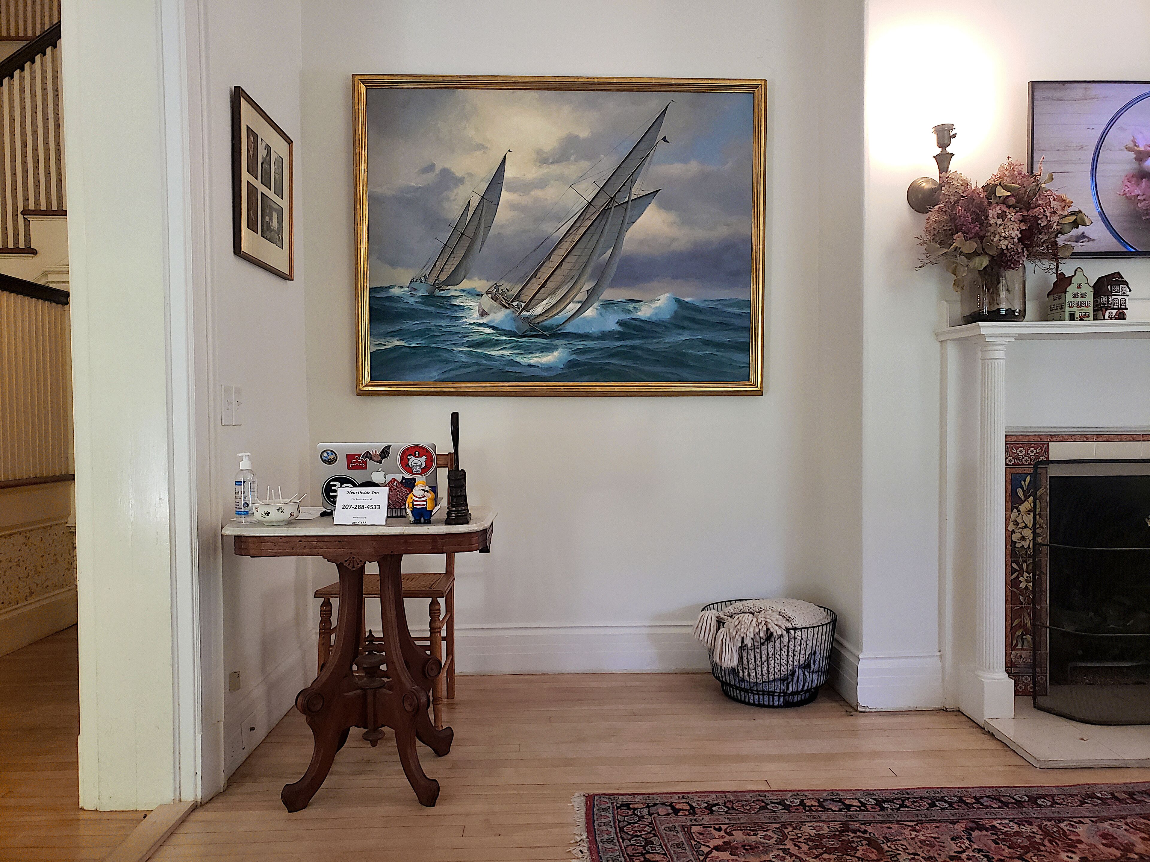 Una habitación con piso de madera, un cuadro de veleros, una mesa con una computadora portátil, un florero con flores y una chimenea.