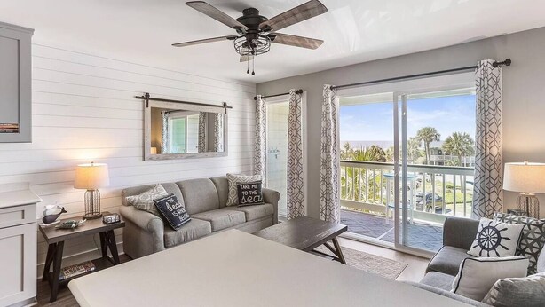 A bedroom with a bed, a desk, a chair, a sofa, a ceiling fan, and a balcony with a view. - Sea Cabin 208 | 1 Bedroom Oceanfront Condo | Pool | Just Renovated (Isle of Palms)