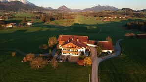 A large, two-story building with a sloped roof, surrounded by green fields and a mountain in the background.