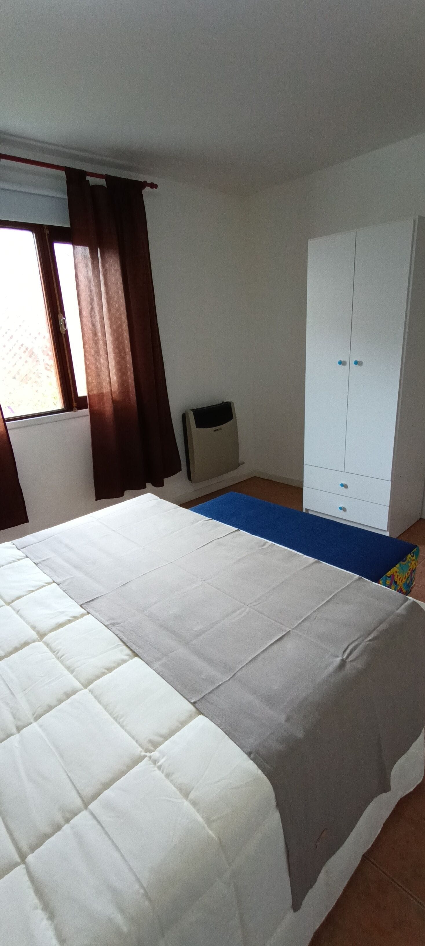 A bedroom with a bed, a wardrobe, a heater, and a window with curtains.