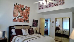 A bedroom with a large bed, a nightstand, a lamp, a ceiling fan, a large floral painting, a mirror, and a door leading to another room. - Panoramic Ocean View Condo (Kihei)