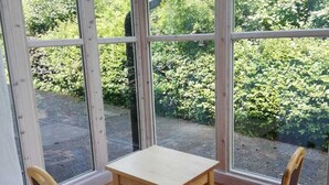 Ein sonnendurchflutetes Zimmer mit einem Holztisch und zwei Stühlen, großen Fenstern und Blick auf Grünfläche.