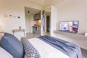 A modern bedroom with a large bed, a TV on a stand, and a view into the kitchen area.