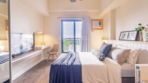 A hotel room with a bed, a desk with a TV, a chair, a window with curtains, and a view of the city. - Astonishing Home Office Balcony Cebu CBD Furnished (Cebu City)