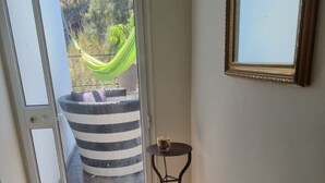 Une pièce dotée d’une porte-fenêtre coulissante qui donne sur un espace extérieur aménagé avec un hamac et un panier à rayures. On y trouve une petite table ronde sur laquelle est posée une tasse, ainsi qu’une chaise bleue dans un coin.