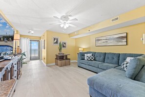 A living room with a blue sofa, a coffee table, and a ceiling fan. There is a door leading to a balcony with a view of the ocean.