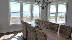 Une salle à manger avec une grande table et des chaises en osier, de grandes fenêtres donnant sur la plage et un lustre noir.