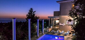 A poolside area with lounge chairs, a pool, and a view of the sea at dusk.