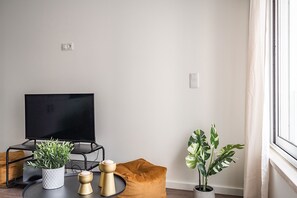 A modern living room with a flat-screen TV, a round coffee table, a brown ottoman, and two potted plants. - Savory Orange Apartment, WiFi, AC !New! (Lisboa)