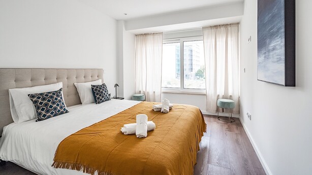 Habitación de hotel con una cama grande, cabecera, ventana con cortinas, mesita de noche y una obra de arte colgada en la pared.