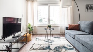 Una sala de estar moderna con televisor de pantalla plana, un sofá gris, una mesita redonda con una planta y un ventanal con cortinas.