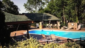 A swimming pool with lounge chairs and a covered area, surrounded by trees and a building with a green roof.
