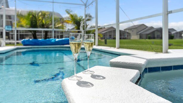 A swimming pool with two glasses of champagne on the edge, surrounded by a white structure and palm trees in the background. - 4BR/3Bath Home, pool/spa, game room, lake view. Closer to ALL ATTRACTIONS (Kissimmee)