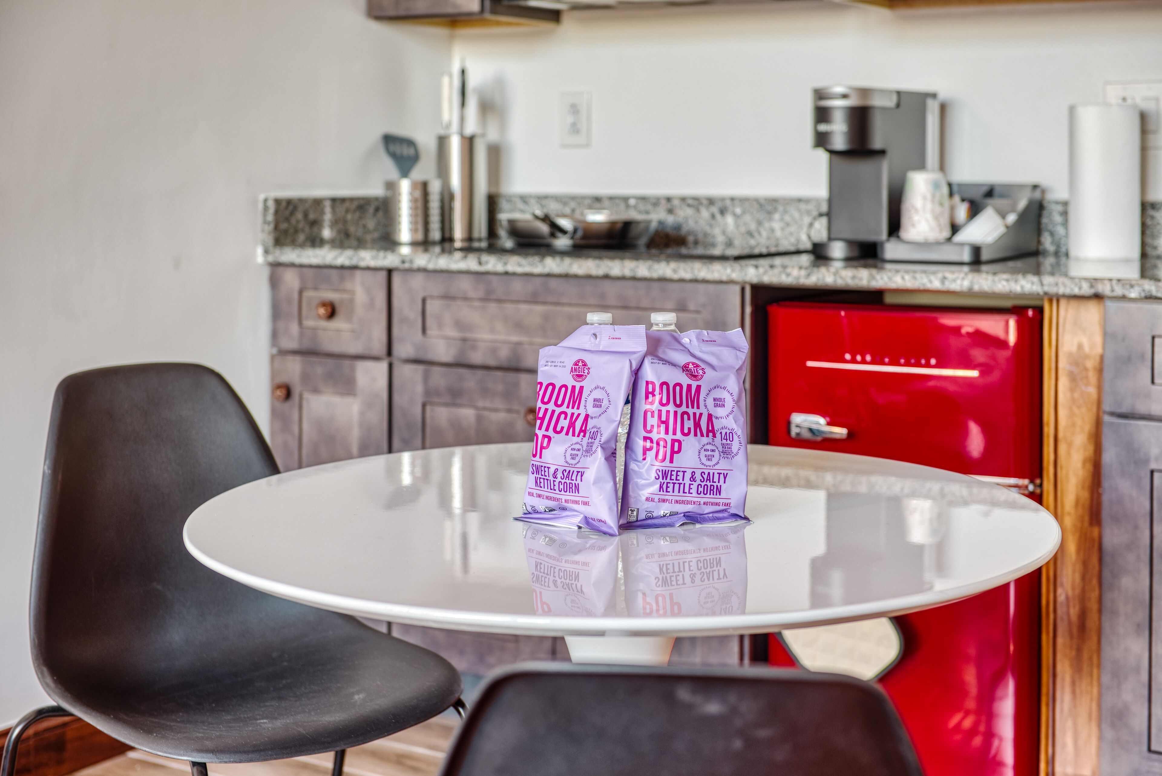 A kitchen with a round table, two chairs, a red appliance, and two bags of popcorn on the table.