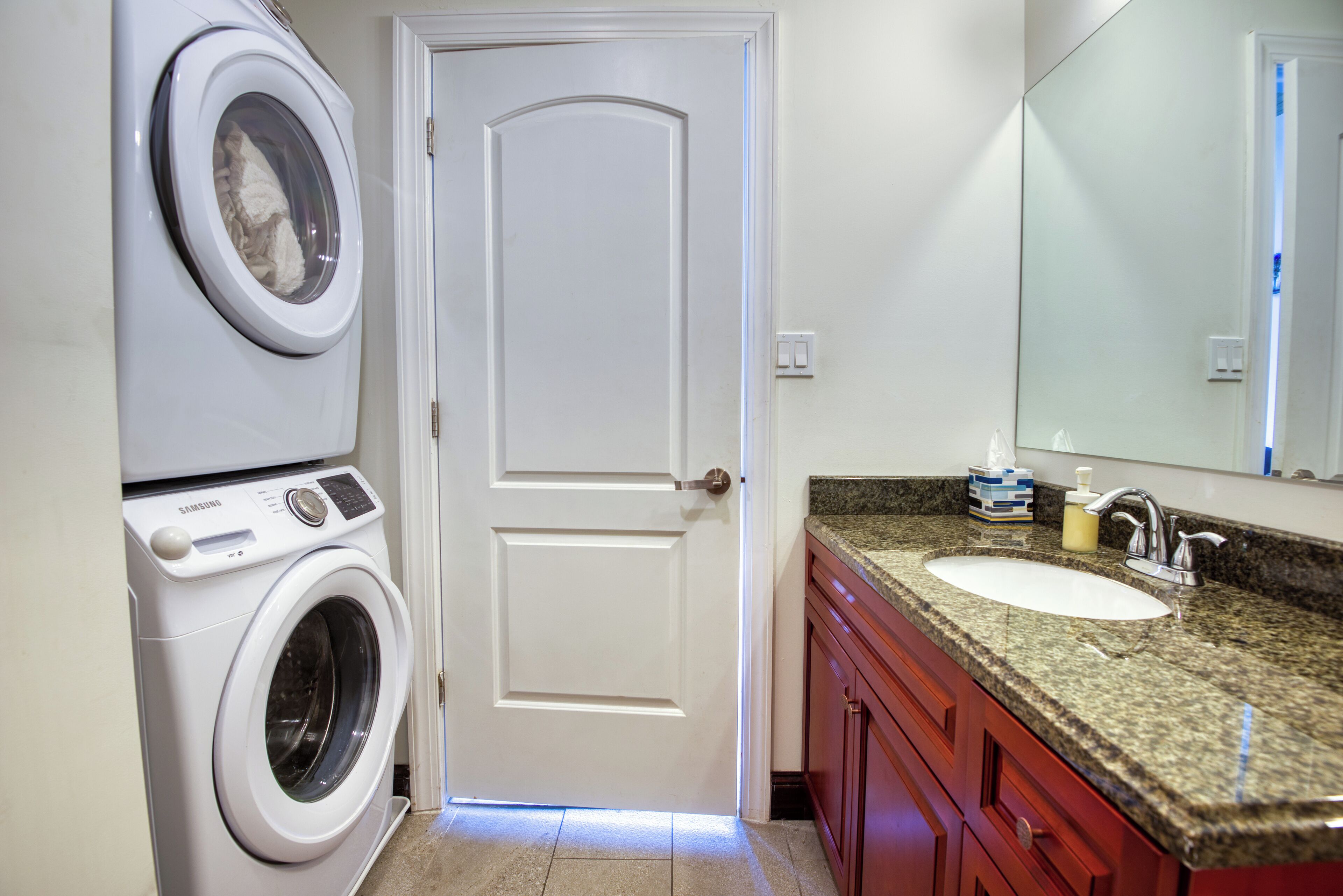 A laundry area with a front-loading washing machine and dryer, a white door, a granite countertop with a sink, and a mirror above the countertop.