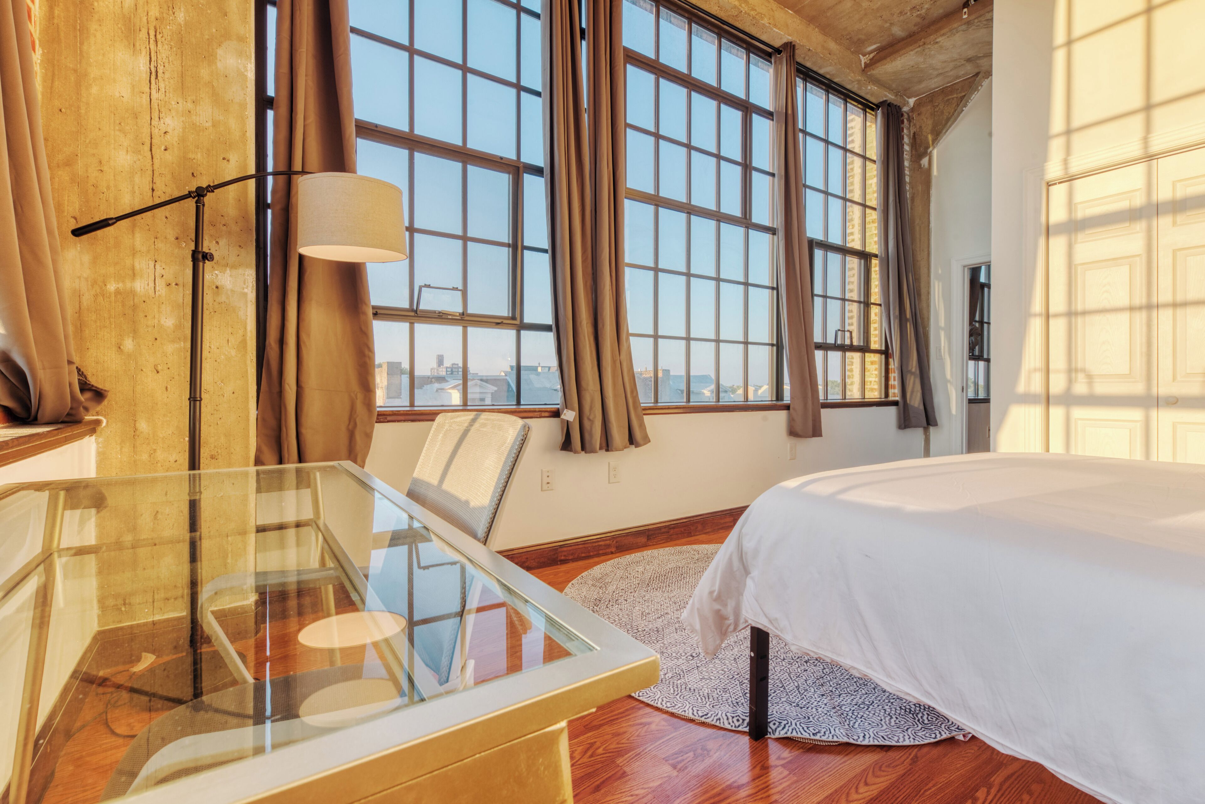 A hotel room with a large window, a bed, a glass table, and a chair.