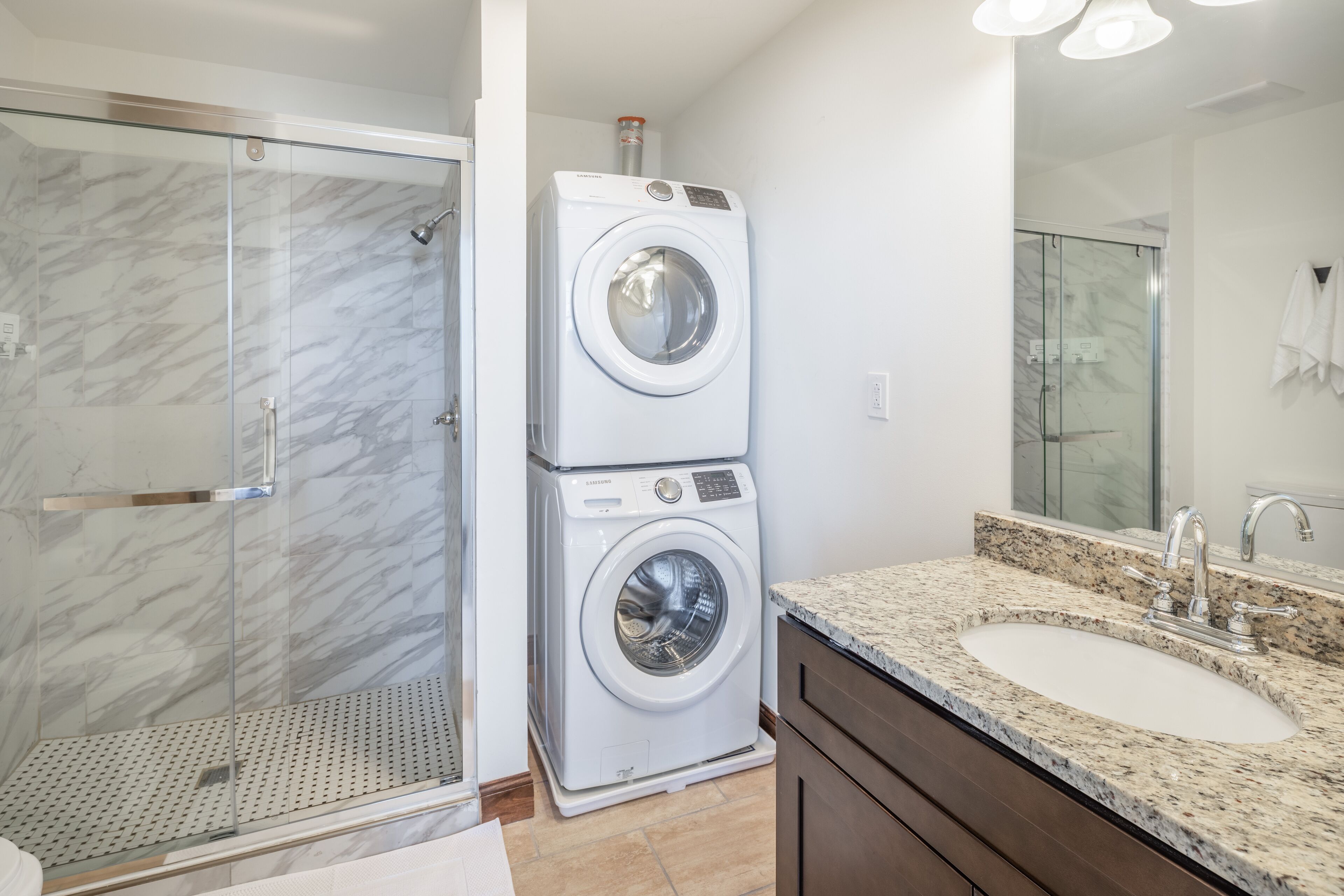 A modern bathroom with a glass-enclosed shower, a stacked washer and dryer, a granite countertop sink, and a large mirror.