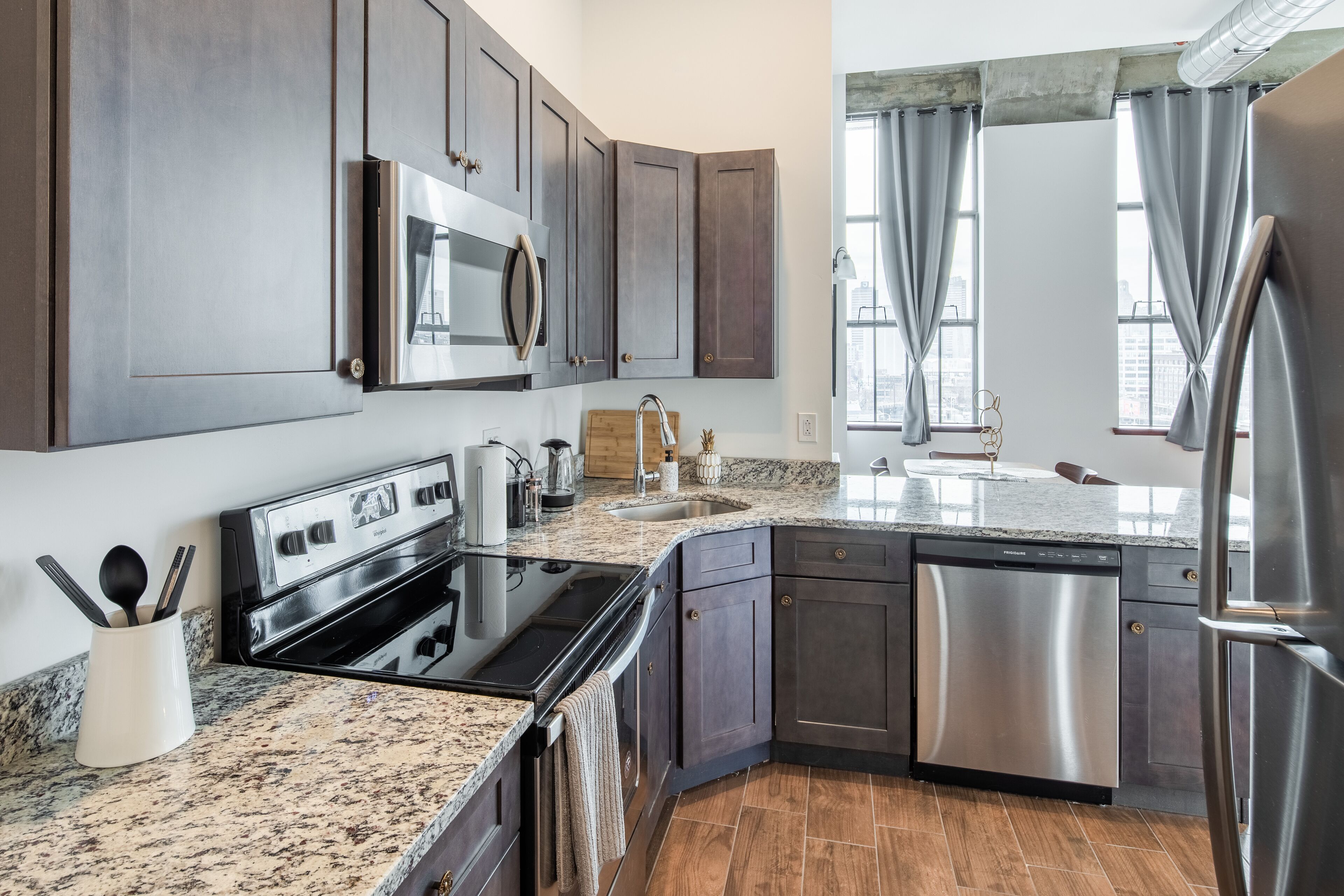 A modern kitchen with dark cabinets, stainless steel appliances, granite countertops, and a large window with curtains.