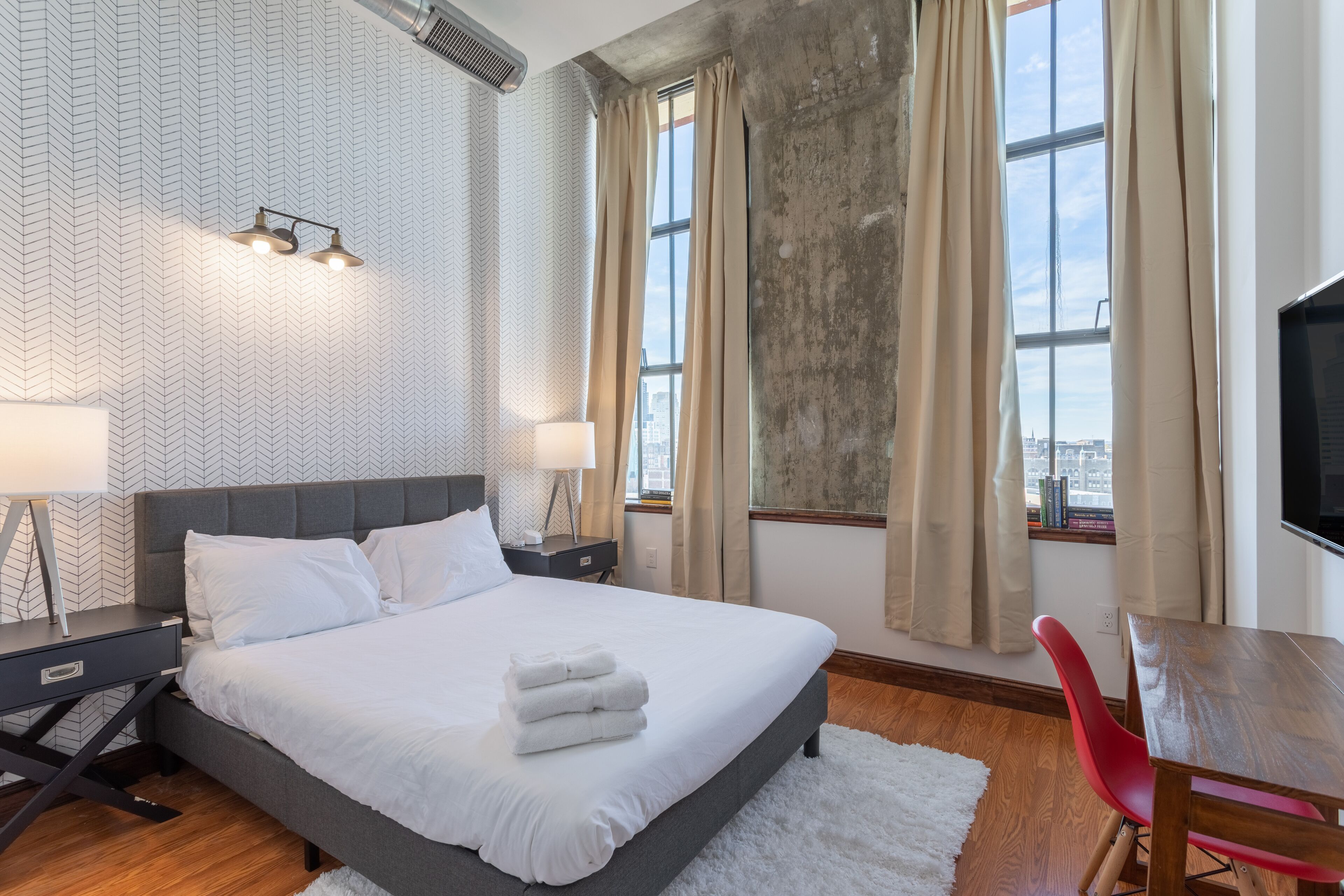 A modern hotel room with a large bed, two bedside tables with lamps, a red chair, a wooden desk, and a large window with curtains.