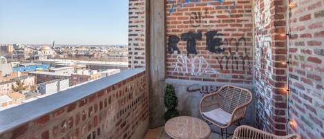 A balcony with brick walls, two wicker chairs, a small table, and a view of a cityscape.