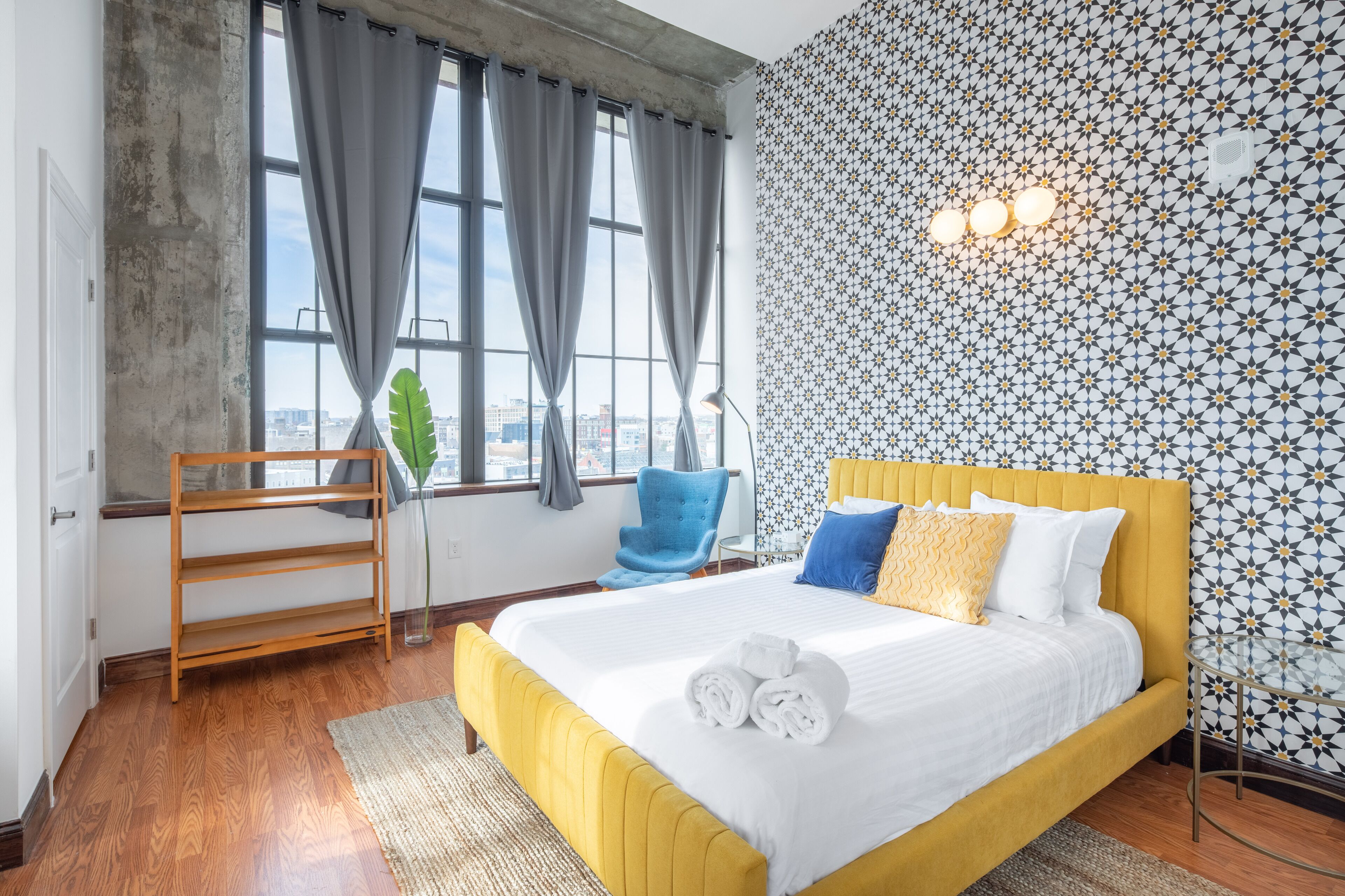 Een hotelkamer met een groot bed, een houten stoel, een bureau en uitzicht op de stad door grote ramen.