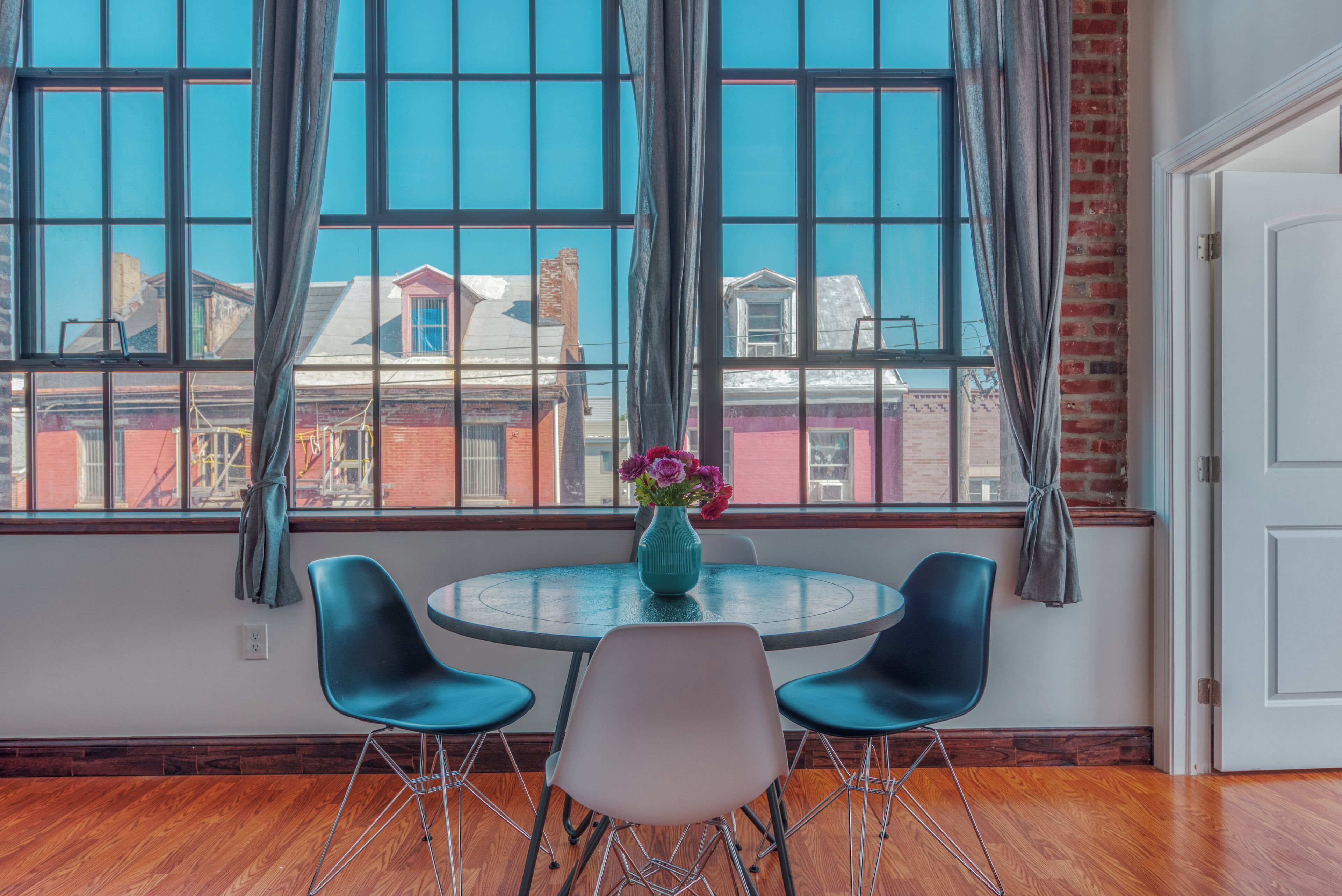 A room with a round table, two blue chairs, a vase with flowers, and a view of buildings through large windows.