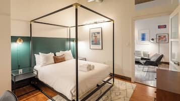 A four-poster bed with white linens and a green headboard, a glass side table, a modern lamp, and a view into a living area with a sofa and a chair.