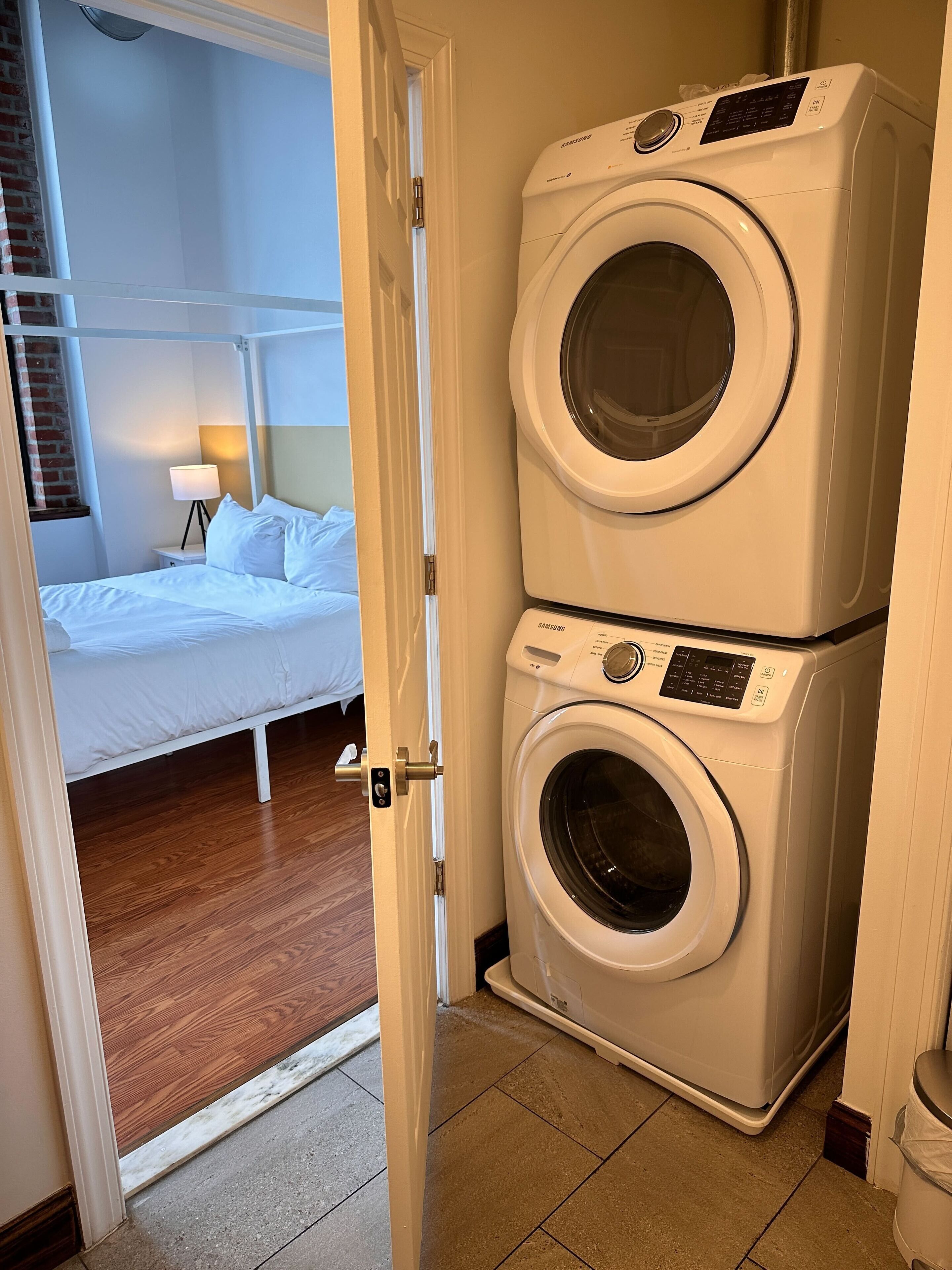 A hotel room with a bed, a nightstand, a lamp, and a washing machine.