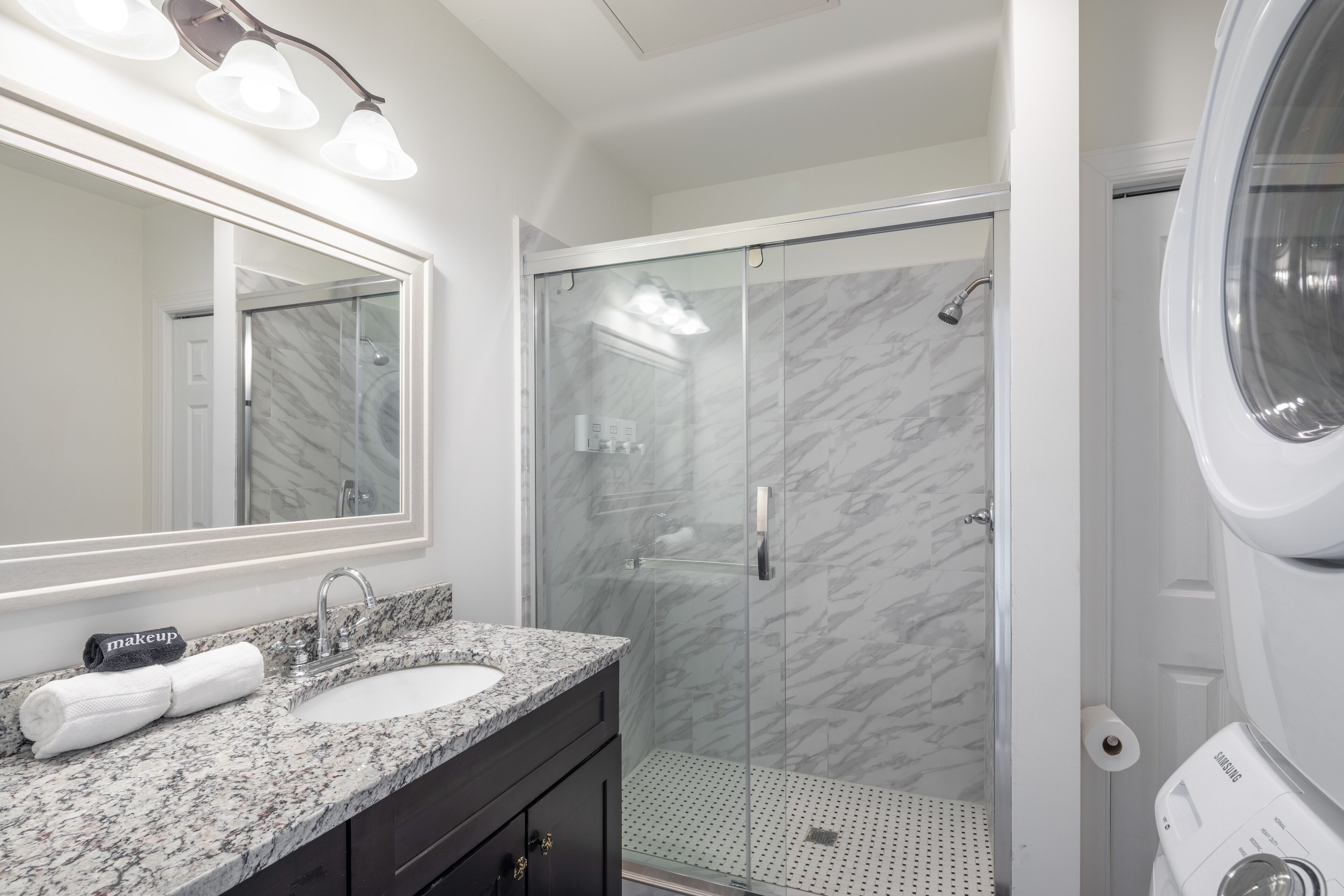 A bathroom with a marble sink, a glass shower enclosure, and a Samsung washing machine.
