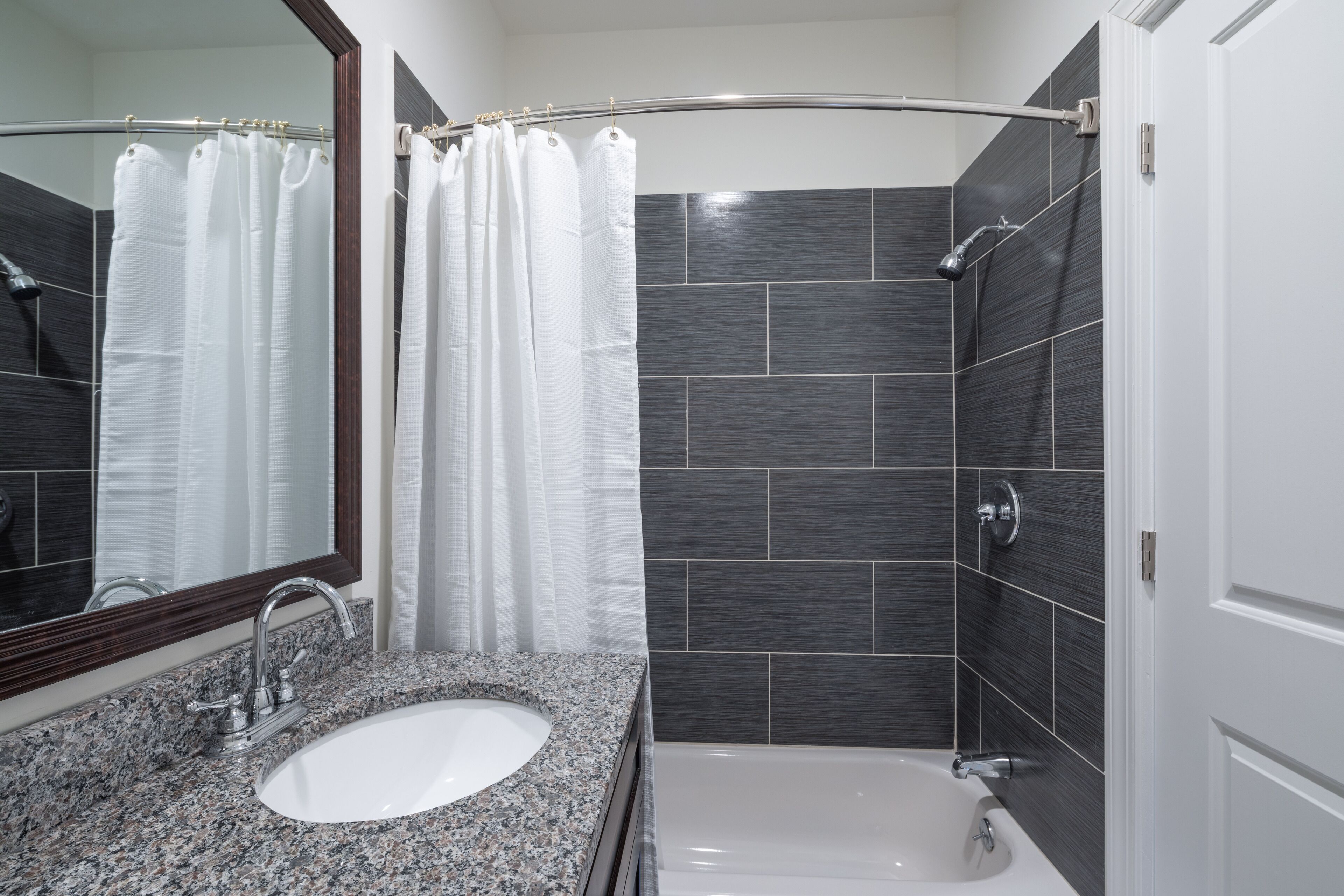 Un baño moderno con lavamanos de granito, una bañera blanca y paredes de azulejos oscuros.