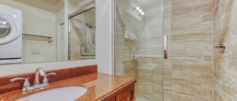 A bathroom with a granite countertop sink, a mirror, a glass-enclosed shower, and a marble wall.