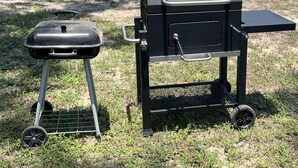 Un barbecue portable et une table de barbecue installés dans un espace extérieur verdoyant, avec un bâtiment bleu en arrière-plan.