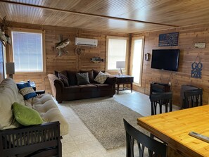 L’intérieur chaleureux d’un chalet avec un plafond en bois, un canapé marron, une table à manger et un téléviseur fixé au mur.