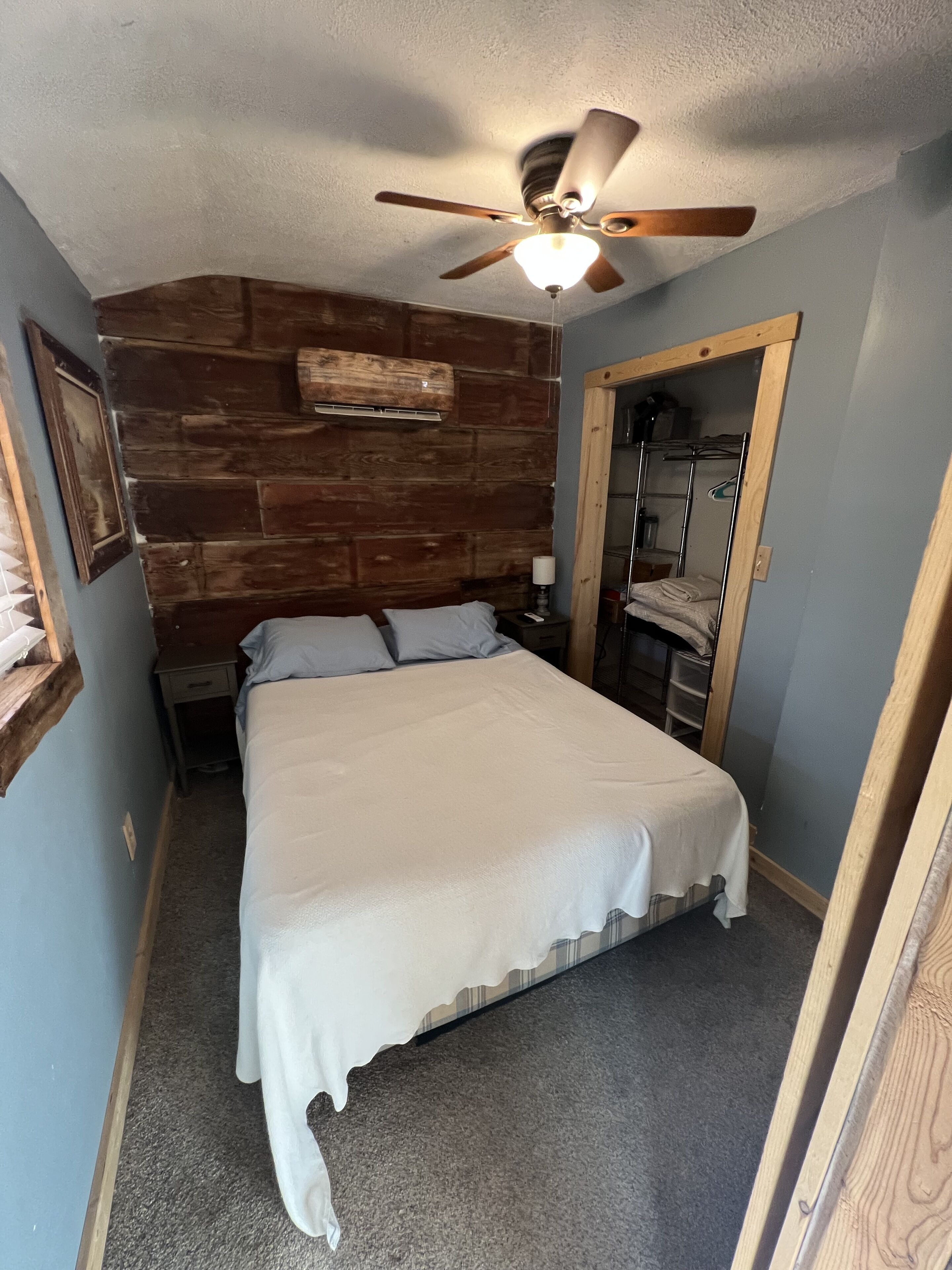 A bedroom with a wooden headboard, a ceiling fan, a bed with white bedding, a nightstand, and a mirror.