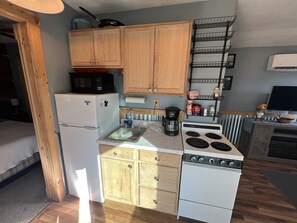 A compact kitchen with wooden cabinets, a refrigerator, a stove, a microwave, a coffee maker, and a shelf with kitchen items.