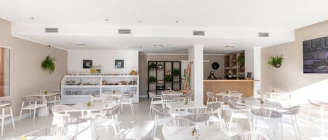 A spacious, well-lit indoor area with multiple white tables and wicker chairs, a reception desk, and shelves with various items.