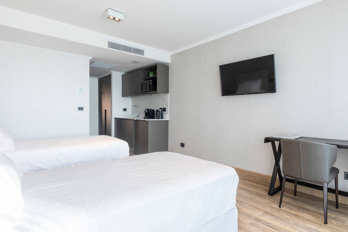 A hotel room with two beds, a desk, a chair, a flat-screen TV, and a kitchenette.