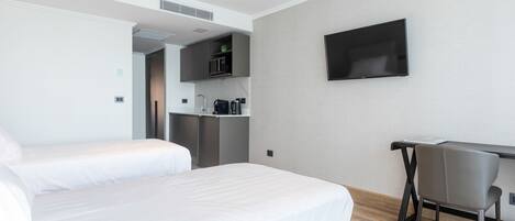 A hotel room with two beds, a desk, a chair, a flat-screen TV, and a kitchenette.