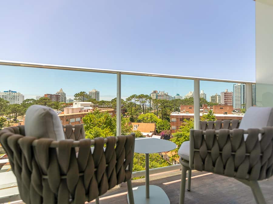 Un balcón con dos sillas y una mesita, que ofrece vistas a un paisaje urbano con edificios y árboles.
