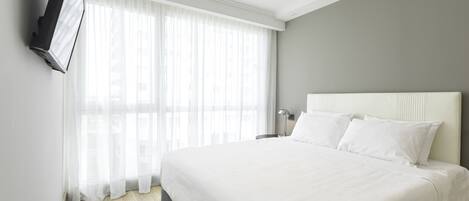 A hotel room with a large bed, a flat-screen TV mounted on the wall, and a window with sheer curtains.