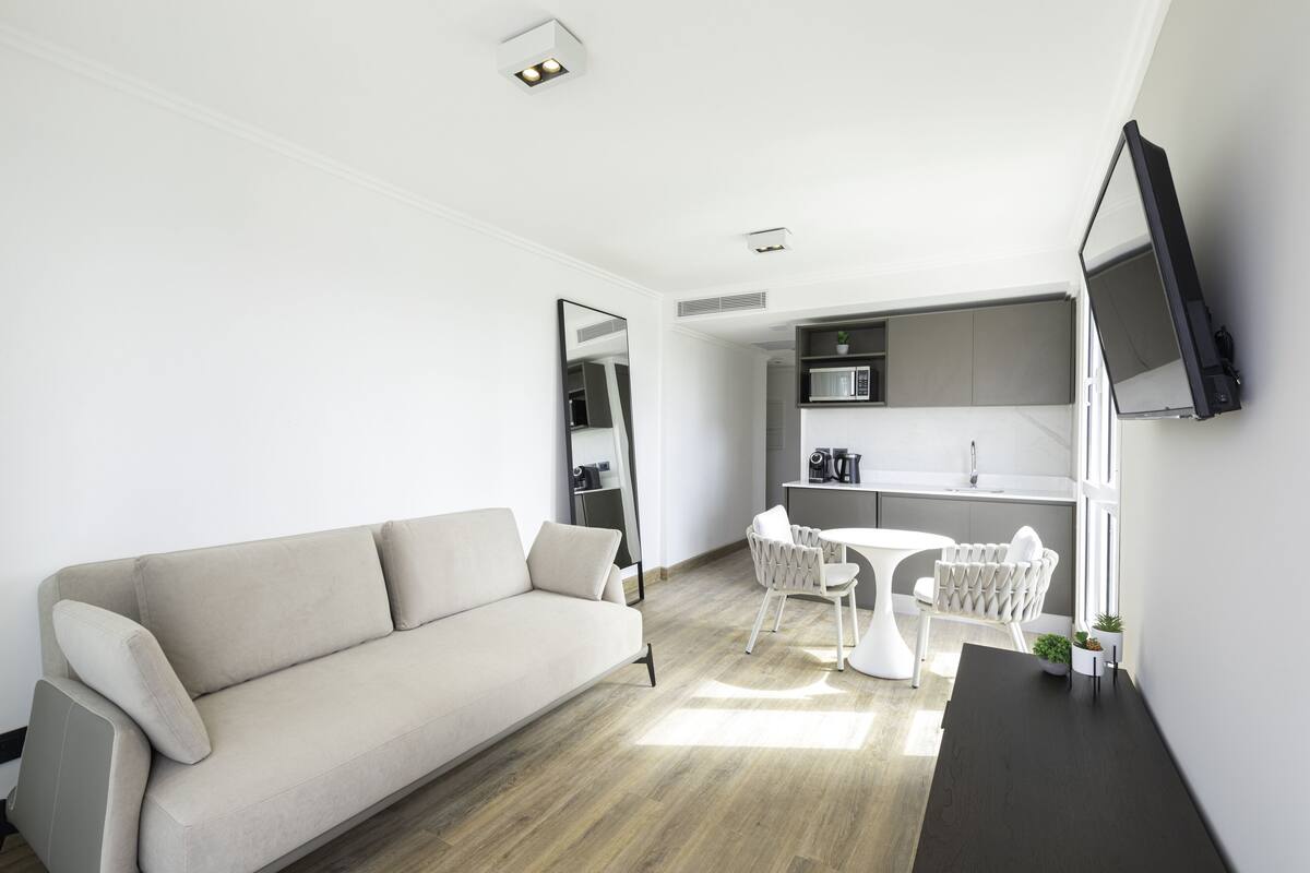 A modern living room with a sofa, a dining table with chairs, a kitchen area with a microwave, and a flat-screen TV mounted on the wall.