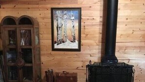 Et rustikt sommerhusinteriør med en trævæg, et indrammet billede af træer, en sort ovn og en pelsfangetæppe på gulvet.