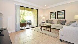 A living room with a sofa, a coffee table, and a TV. There is a sliding glass door leading to an outdoor area with a table and chairs.