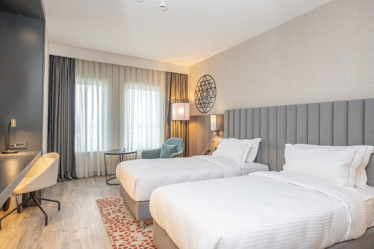 A hotel room with two beds, a desk, a chair, a small table, and a decorative wall piece.