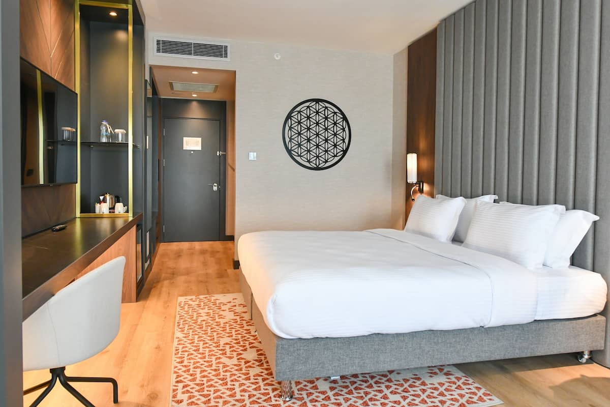 A modern hotel room with a large bed, a desk with a chair, a wardrobe, and a decorative wall piece.