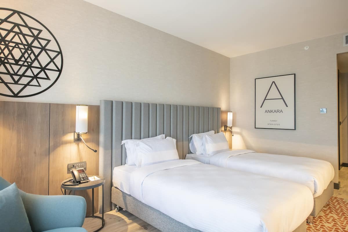 A hotel room with two beds, a small desk, a chair, a phone, and a wall-mounted artwork.