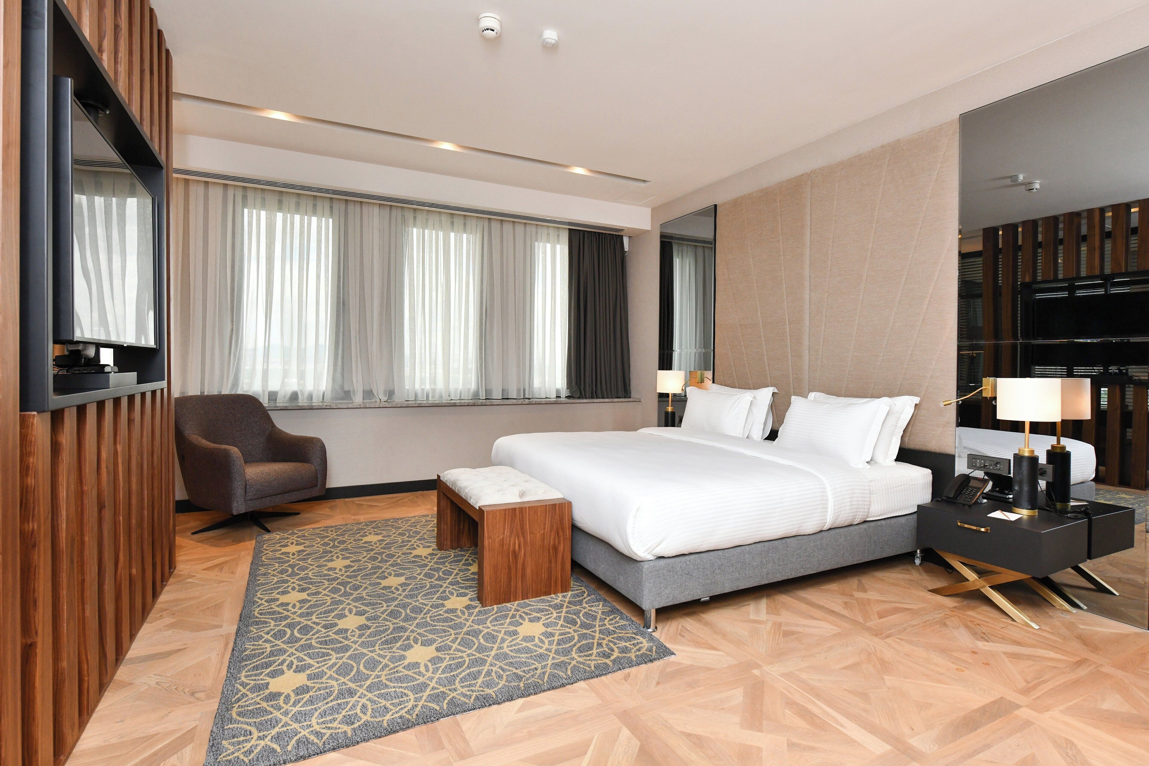 Una habitación de hotel moderna con una cama grande, una zona de estar con una silla, una mesita y un televisor montado en la pared, además de un ventanal con cortinas.