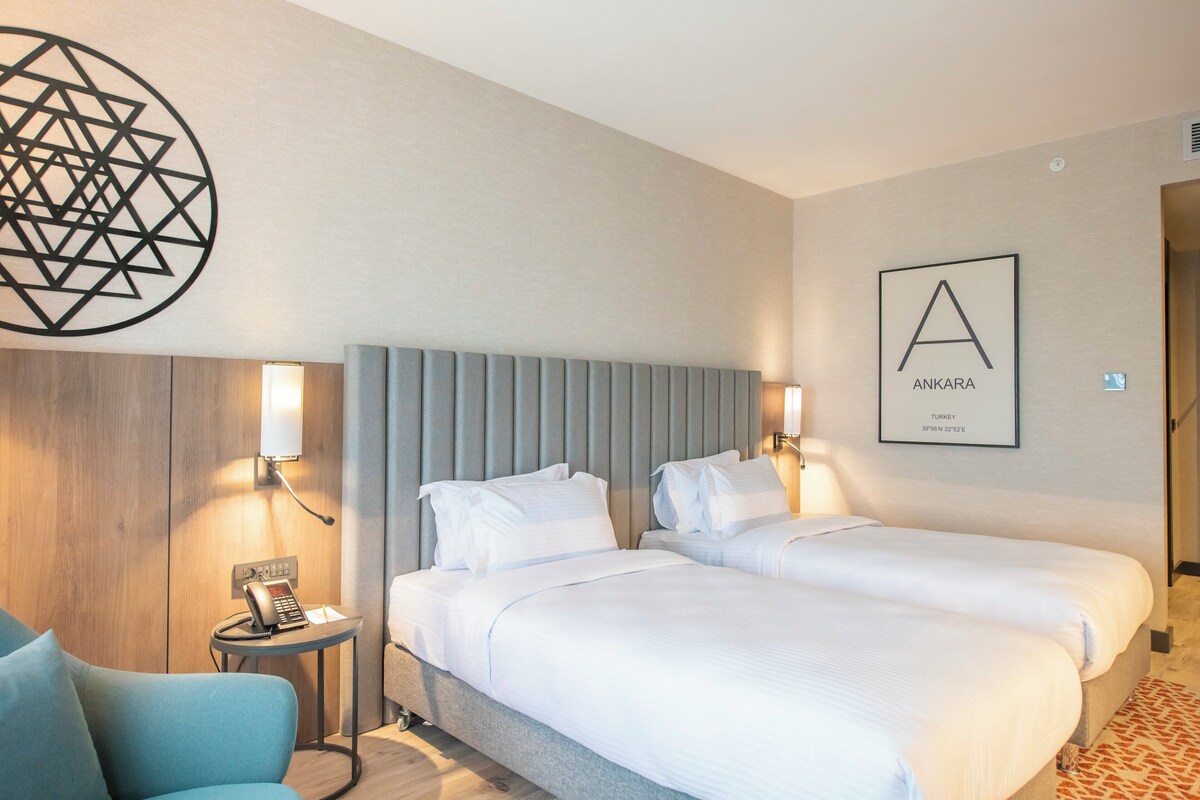 A hotel room with two beds, a small desk, a chair, a phone, and a wall-mounted artwork.